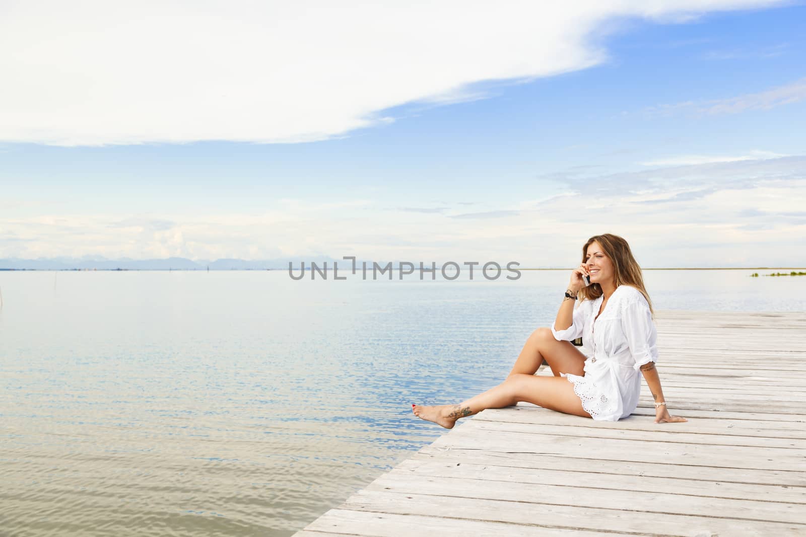Smiling beautiful young woman sitting on a pier and talking on t by alessandroguerriero