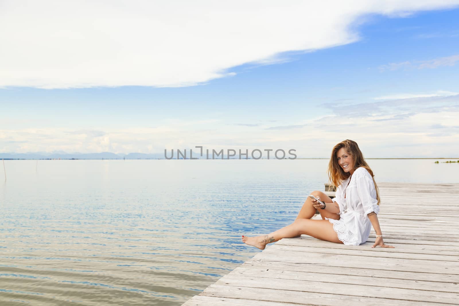 Smiling beautiful young woman sitting on a pier and using a mobi by alessandroguerriero