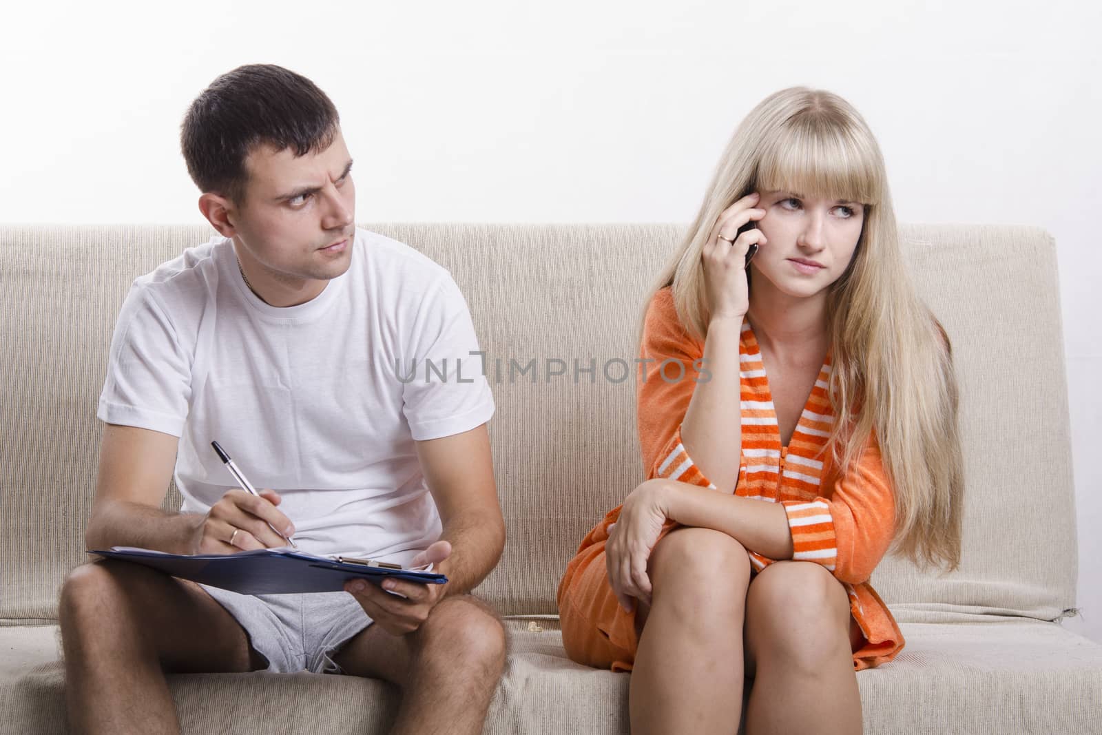 Young couple sitting on the couch. Girl on the phone, the guy sitting with the directory and writes a note of the telephone conversation. He is very surprised to hear, girl concerned. The pair dressed home