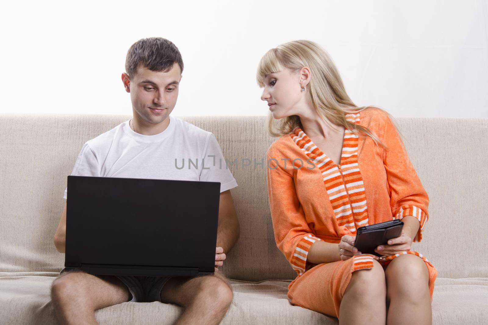 A boy and a girl sitting on the couch. A guy sitting with a laptop, a girl with a tablet. The guy saw something interesting, the girl looked with interest at his laptop.