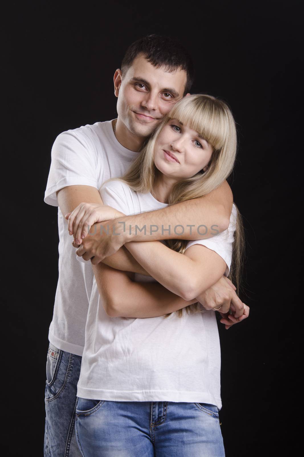 A half-length portrait of a young couple. Guy hugging a girl back. Studio, black background.