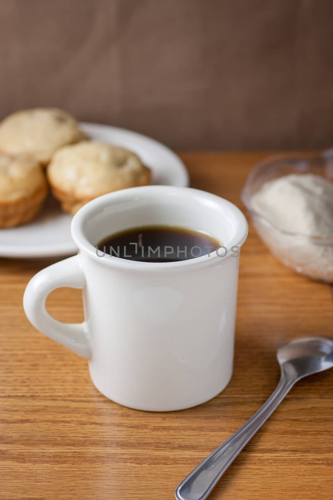 A cup of coffee and metal spoon on a wooden table with apple muffins and a sugar bowl in the background.