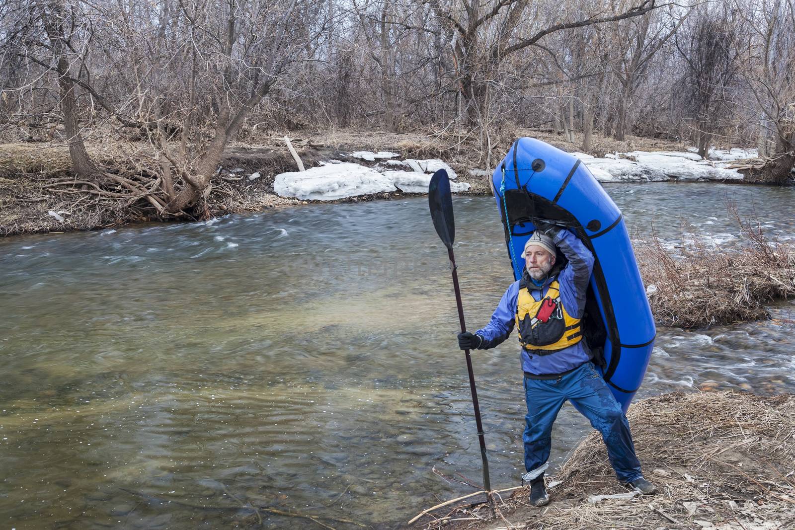 senior male carrying a packraft (one-person light raft used for expedition or adventure racing) on the shore of Cache la Poudre River in Fort Collins, Colorado, winter or early spring scenery