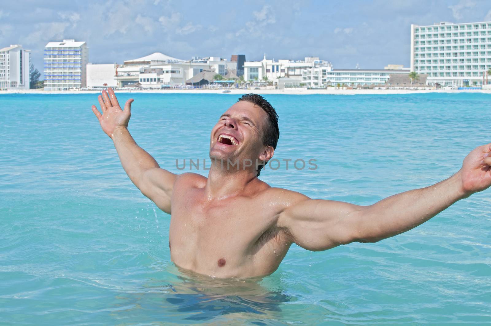 Young fit and shirtless Caucasian man celebrates with arms outstretched in shallow tropical waters of beach resort