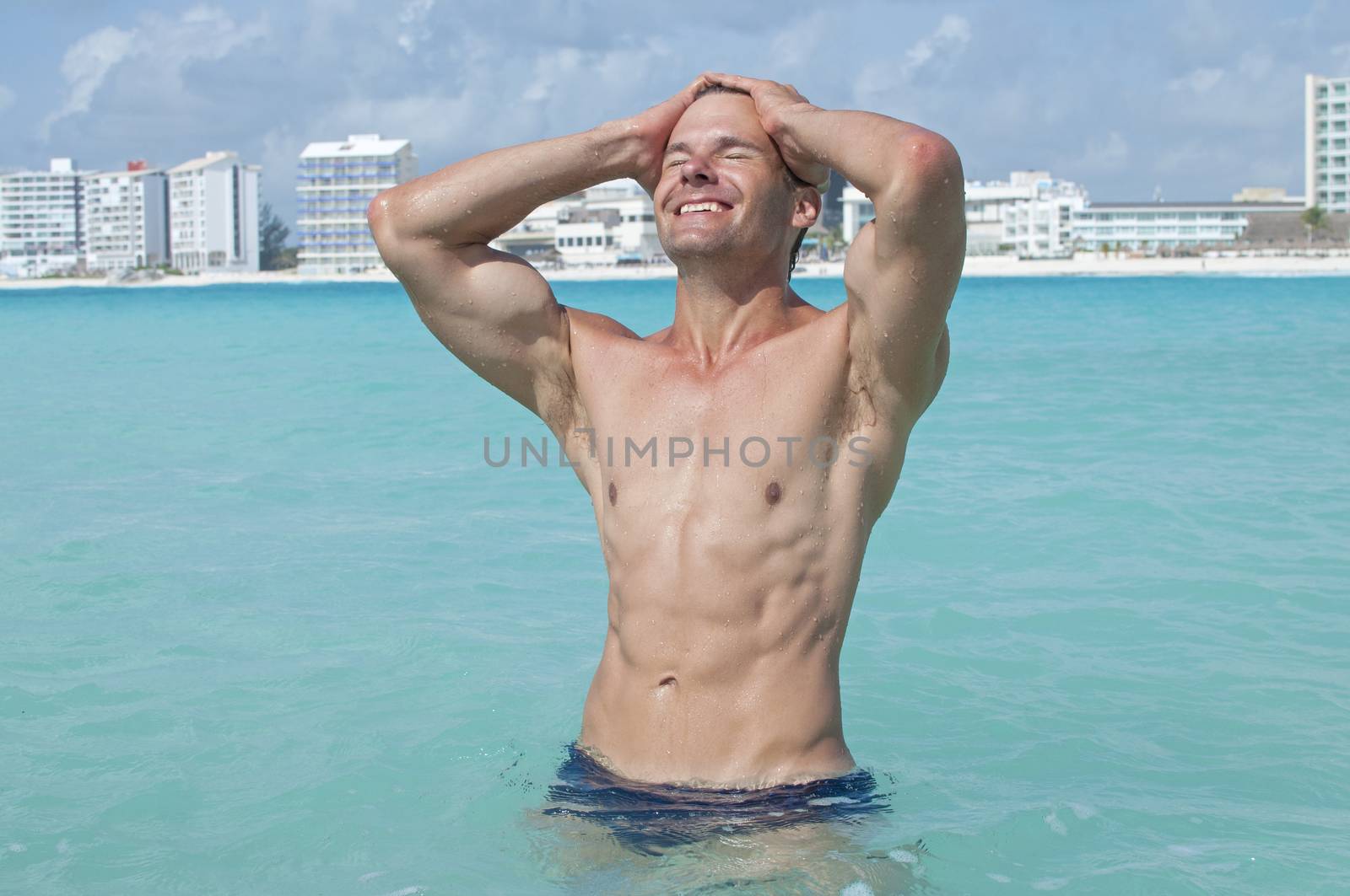 Lean muscular shirtless Caucasian man with abs smiles and pulls hair back while standing in beautiful shallow tropical sea with large resorts in background