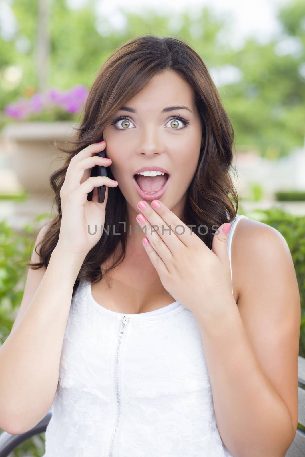 Shocked Young Adult Female Talking on Cell Phone Outdoors on Bench.