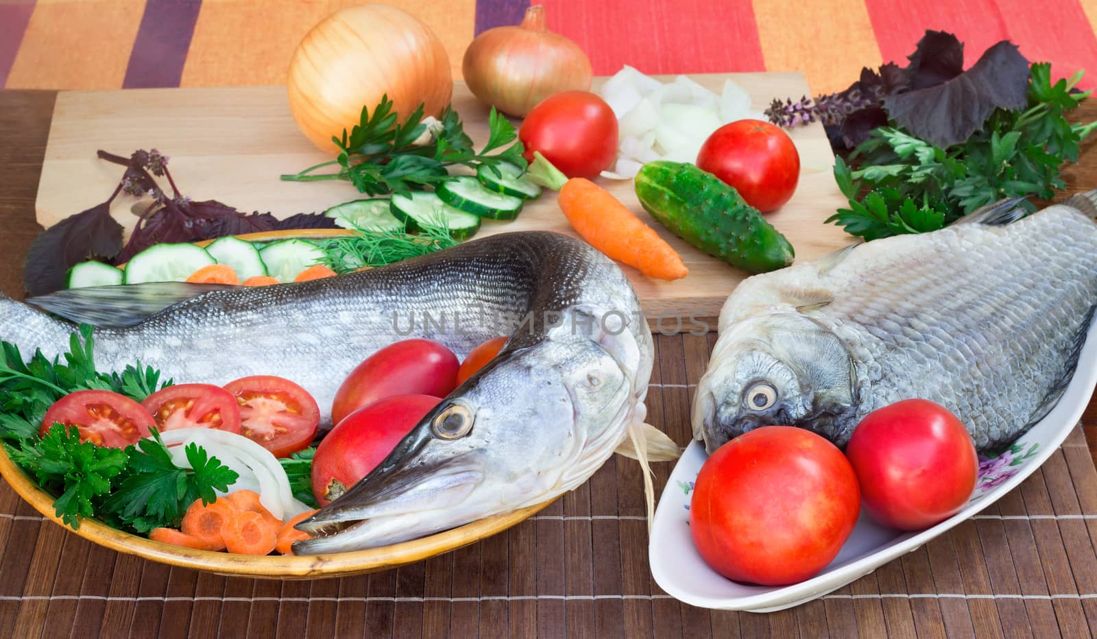 On a table on a chopping board there are a fish, cucumbers, tomatoes, onions, spices and parsley greens.