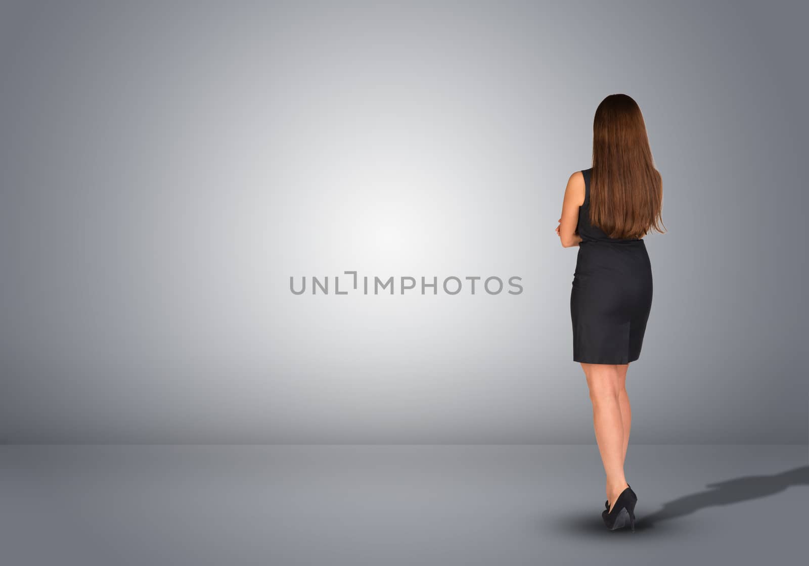 Businesswoman in dress standing in an empty gray room. Rear view. Hands crossed on his chest