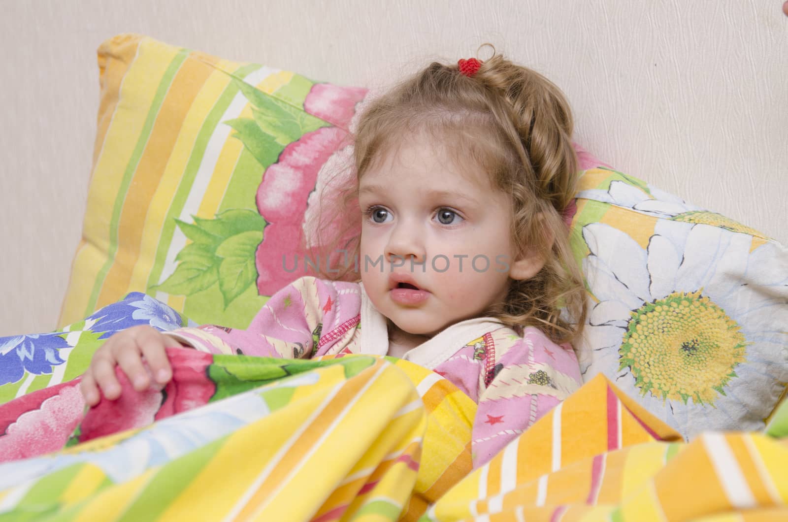 Two-year-old girl is in bed. Pillow put to the wall and covered her bright blanket. Home furnishings.