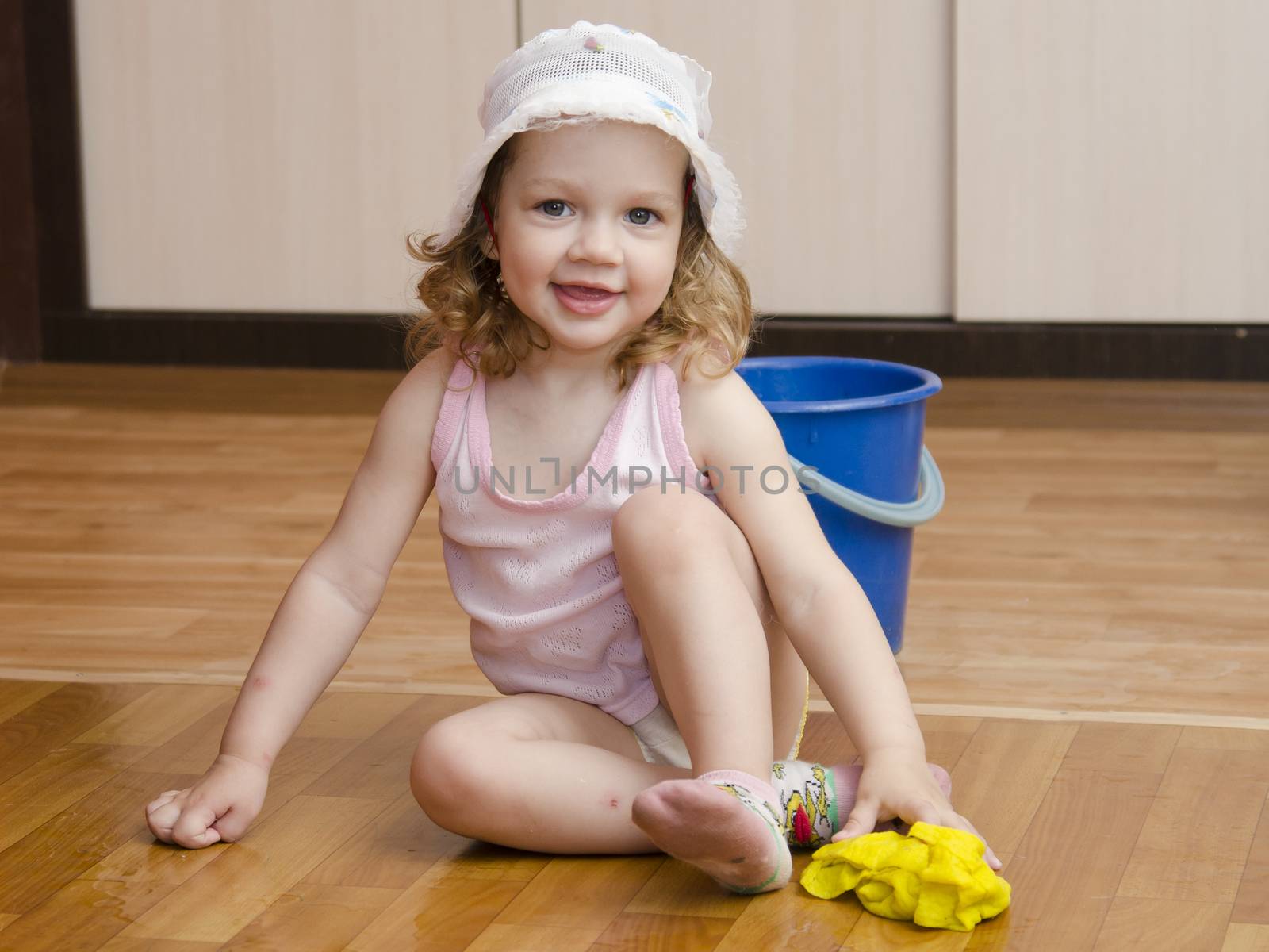 Two-year-old girl with pleasure and fun washing the floors in the room. Nearby is a small blue bucket