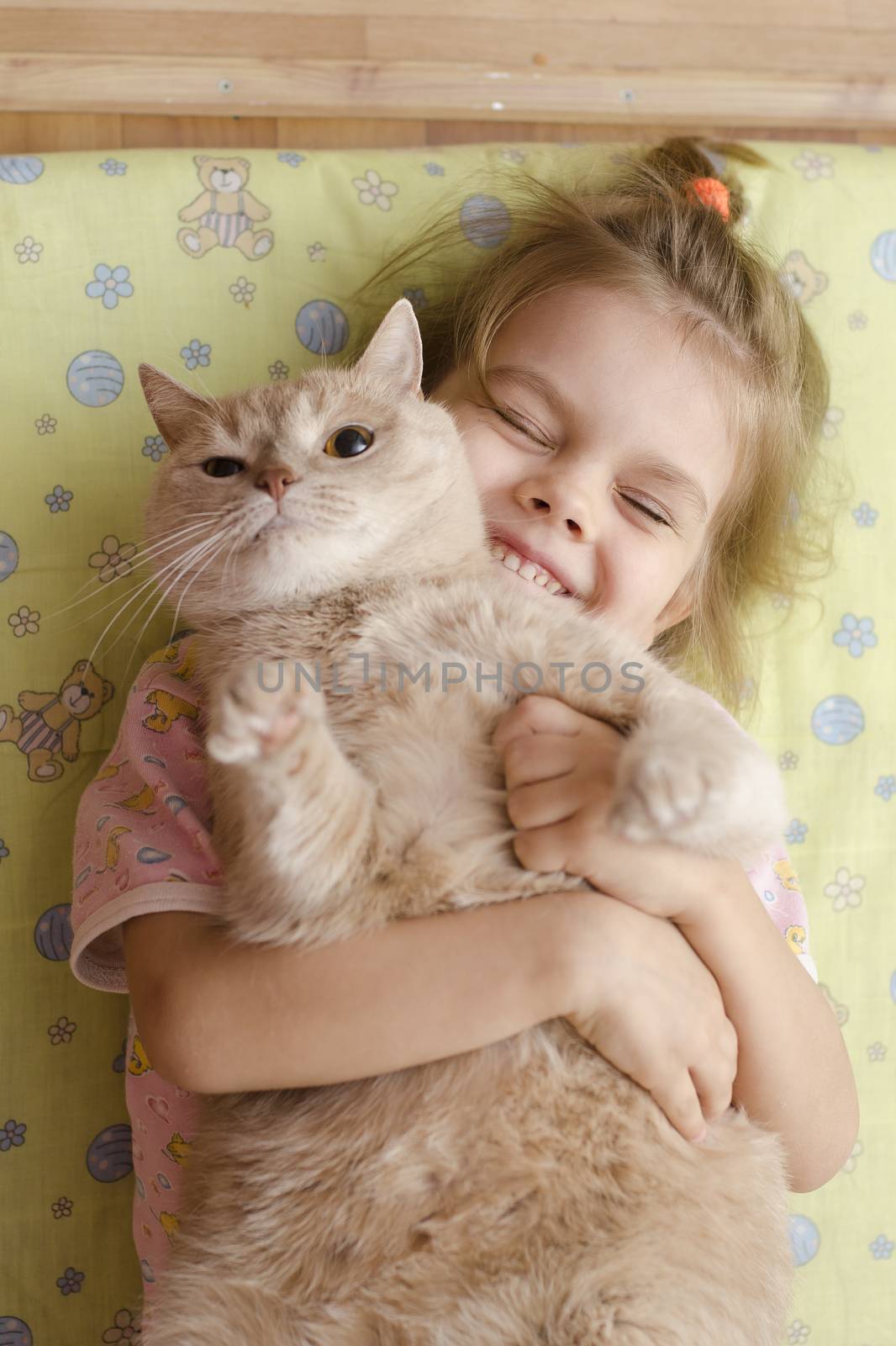 The little girl hugging the cat lying on a mattress on the floor. The girl smiles. He tries to escape