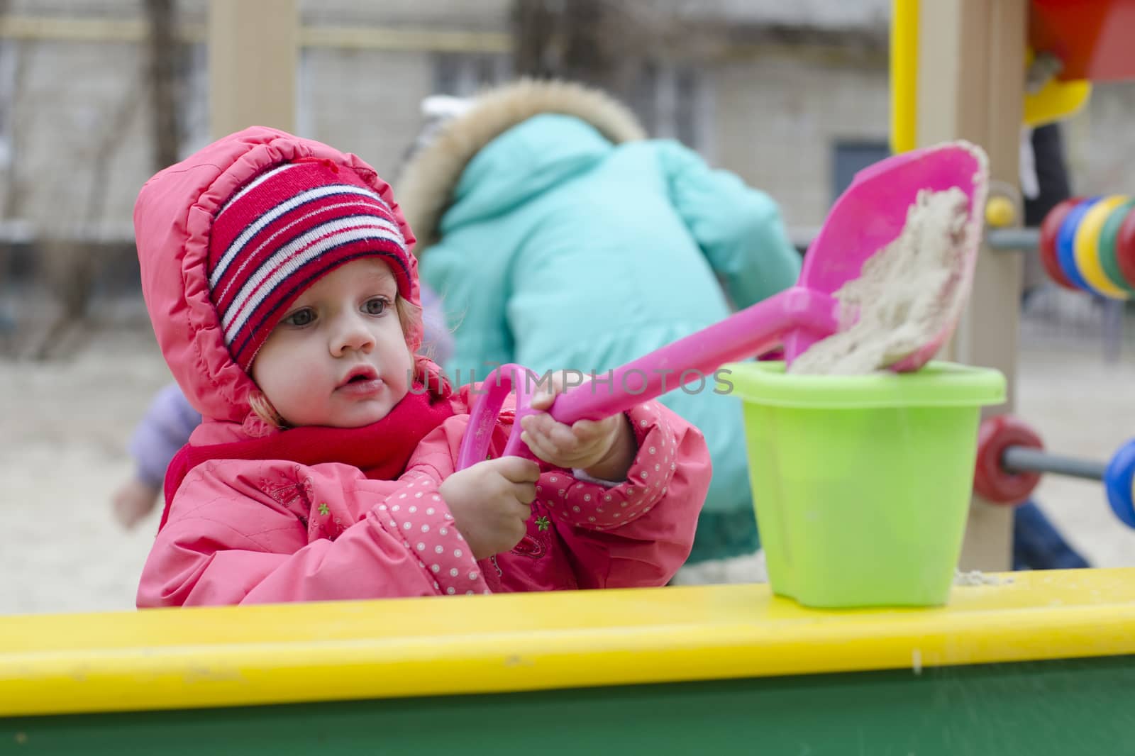 The little girl pours sand in the bucket, while in the sandbox. In early spring. Girl dressed in warm clothes