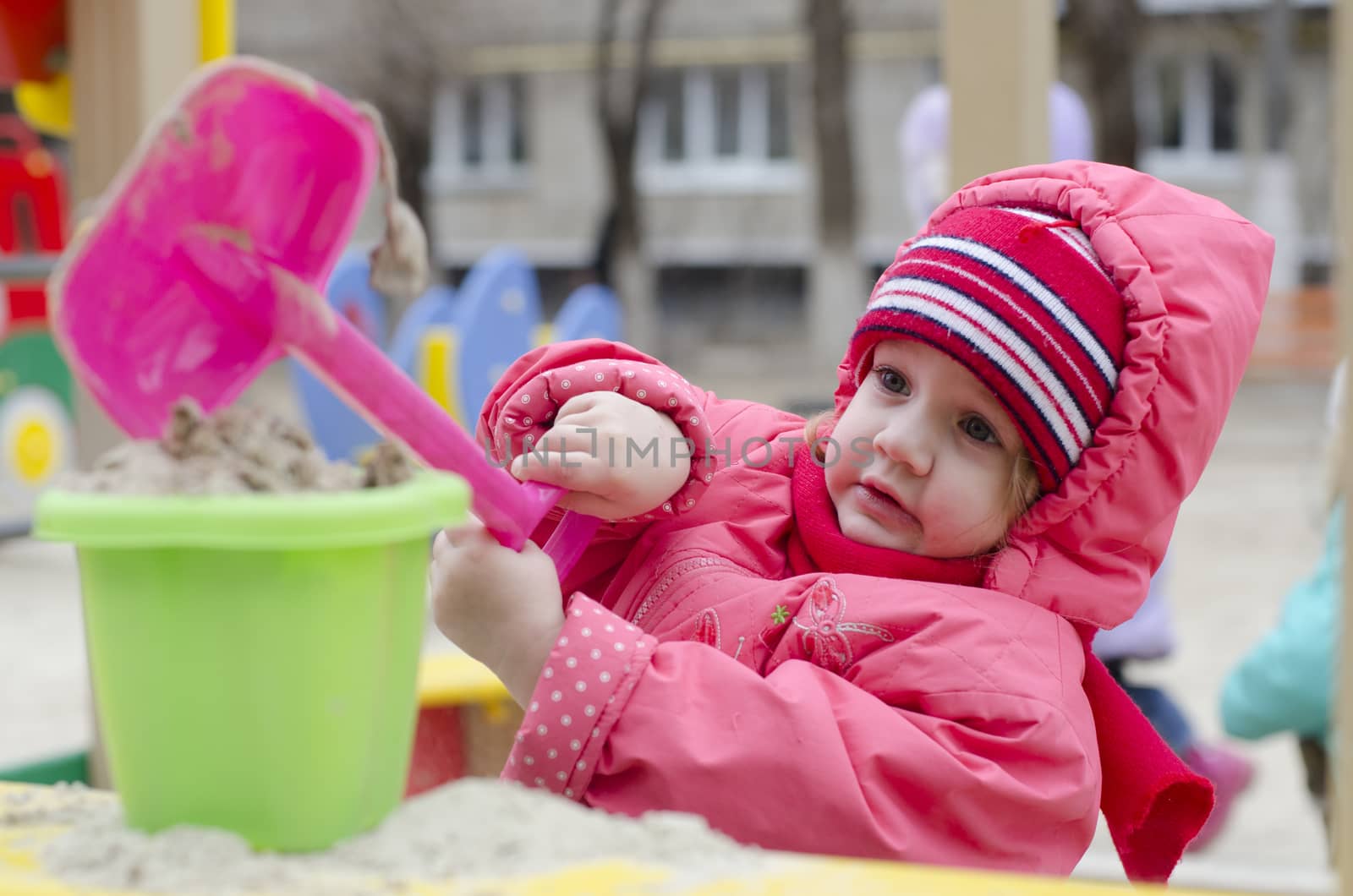 The little girl pours sand in the bucket, while in the sandbox. In early spring. Girl dressed in warm clothes