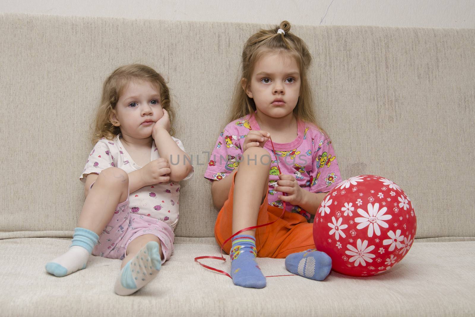 Two little girls sit on the couch and watch TV. Lies next to the ball