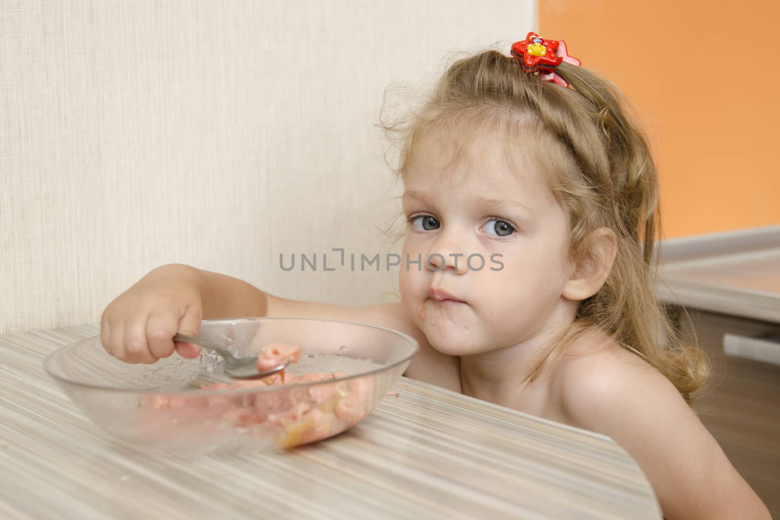 The child sits in the kitchen and eats porridge at the table. The child is distracted and with views of the inquiring looks into the frame.