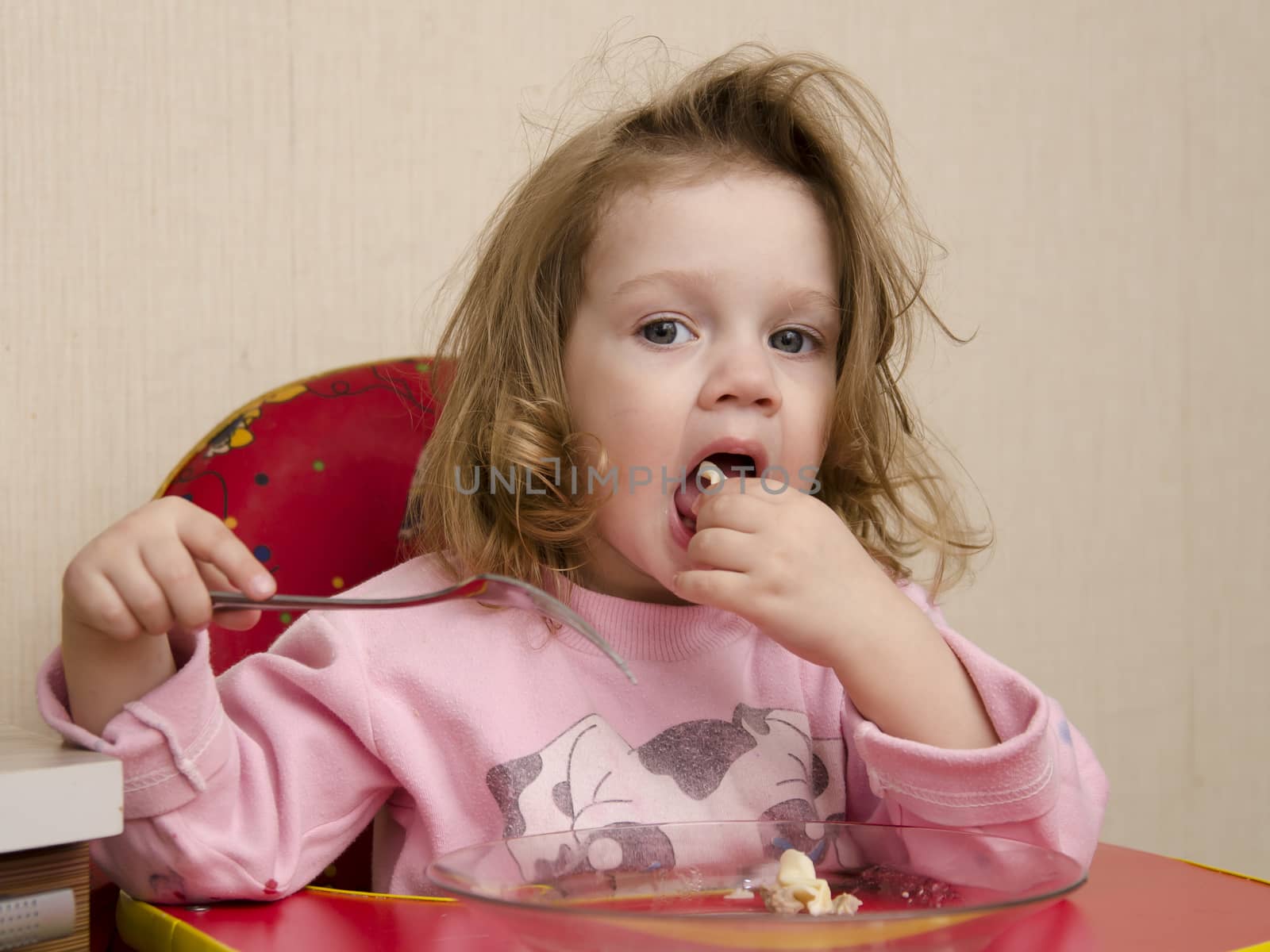Two-year-old girl eats sitting at the table in the kitchen. Girl eats with a fork.