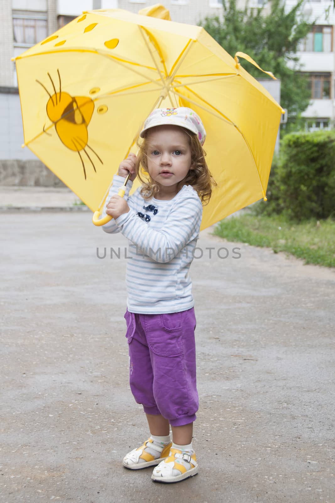 Merry little girl stands with the yellow umbrella on an asphalt road on the street