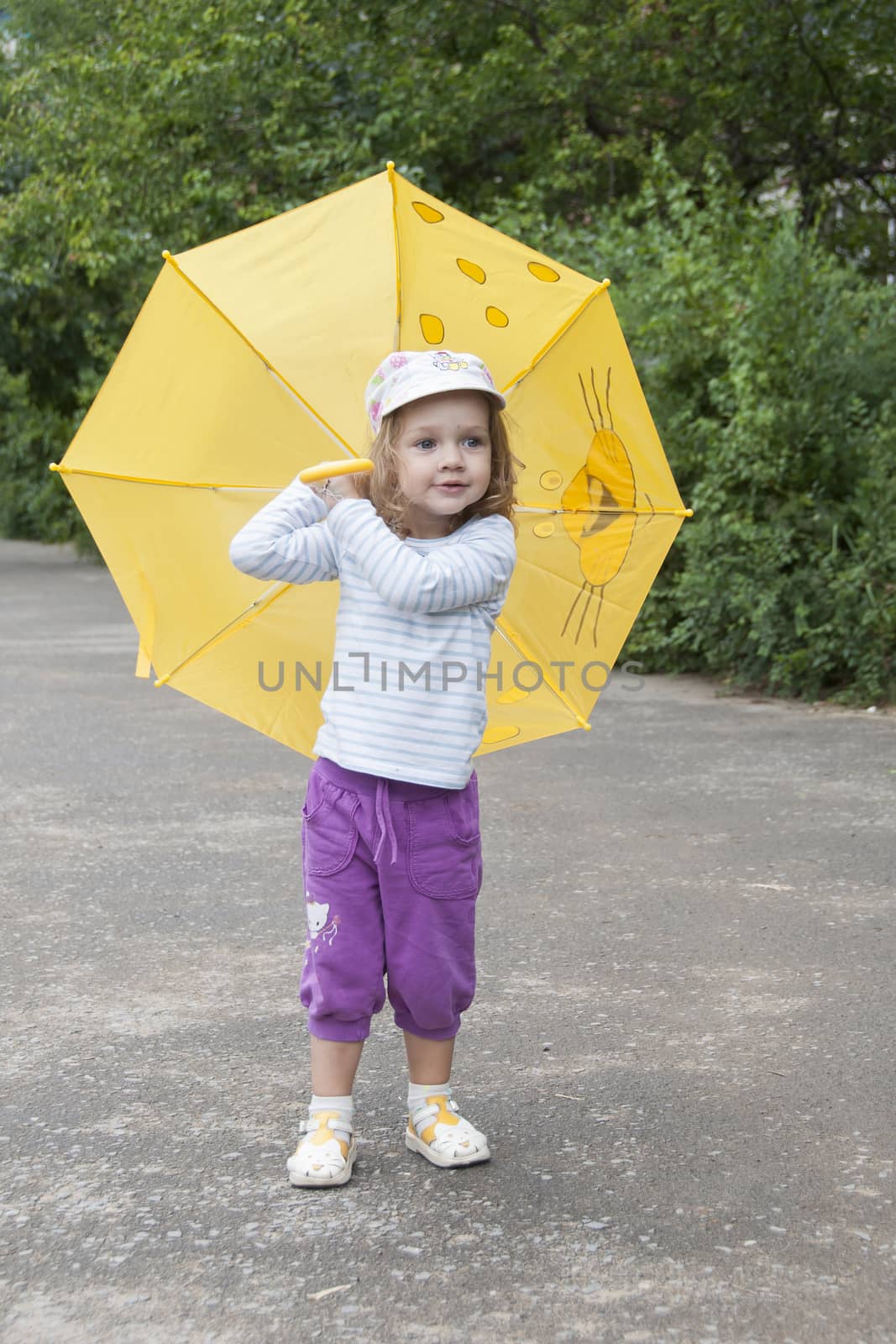 Merry little girl stands with the yellow umbrella on an asphalt road on the street