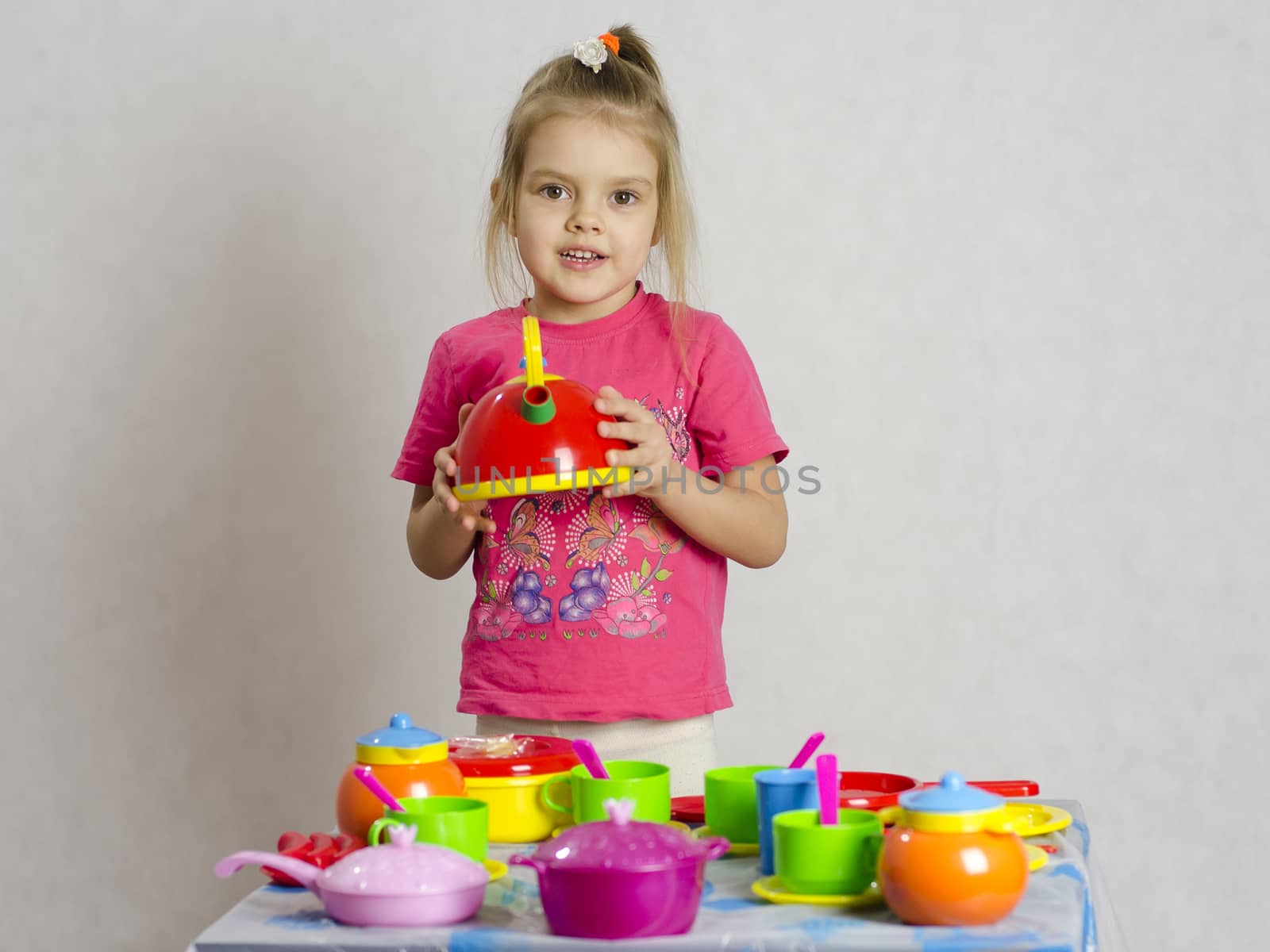 Girl plays child kitchen utensils. She picked up the teapot and looks in the frame. A light background, a Studio.