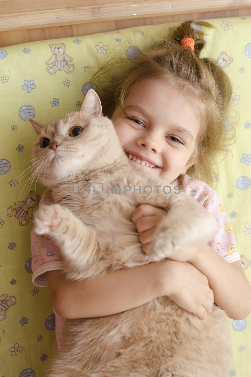 The little girl hugging the cat lying on a mattress on the floor. The girl smiles. He tries to escape