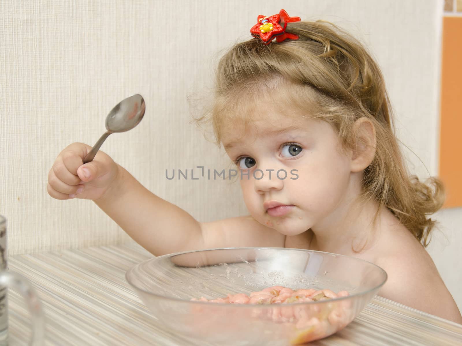 The child sits in the kitchen and eats porridge at the table. The child is distracted and with views of the inquiring looks into the frame.