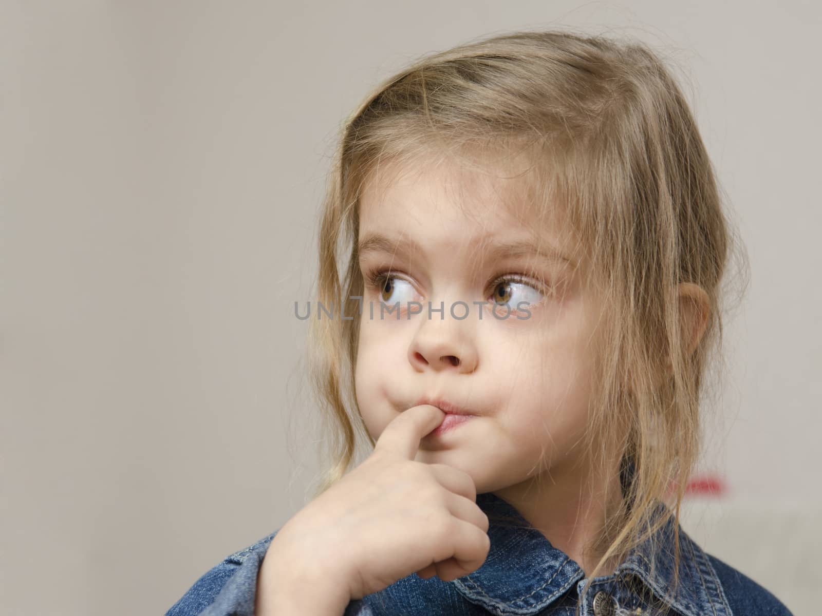Four-year-old girl stuck her index finger in her mouth and enthusiastically looking left. Girl dressed in a denim jacket. Portrait
