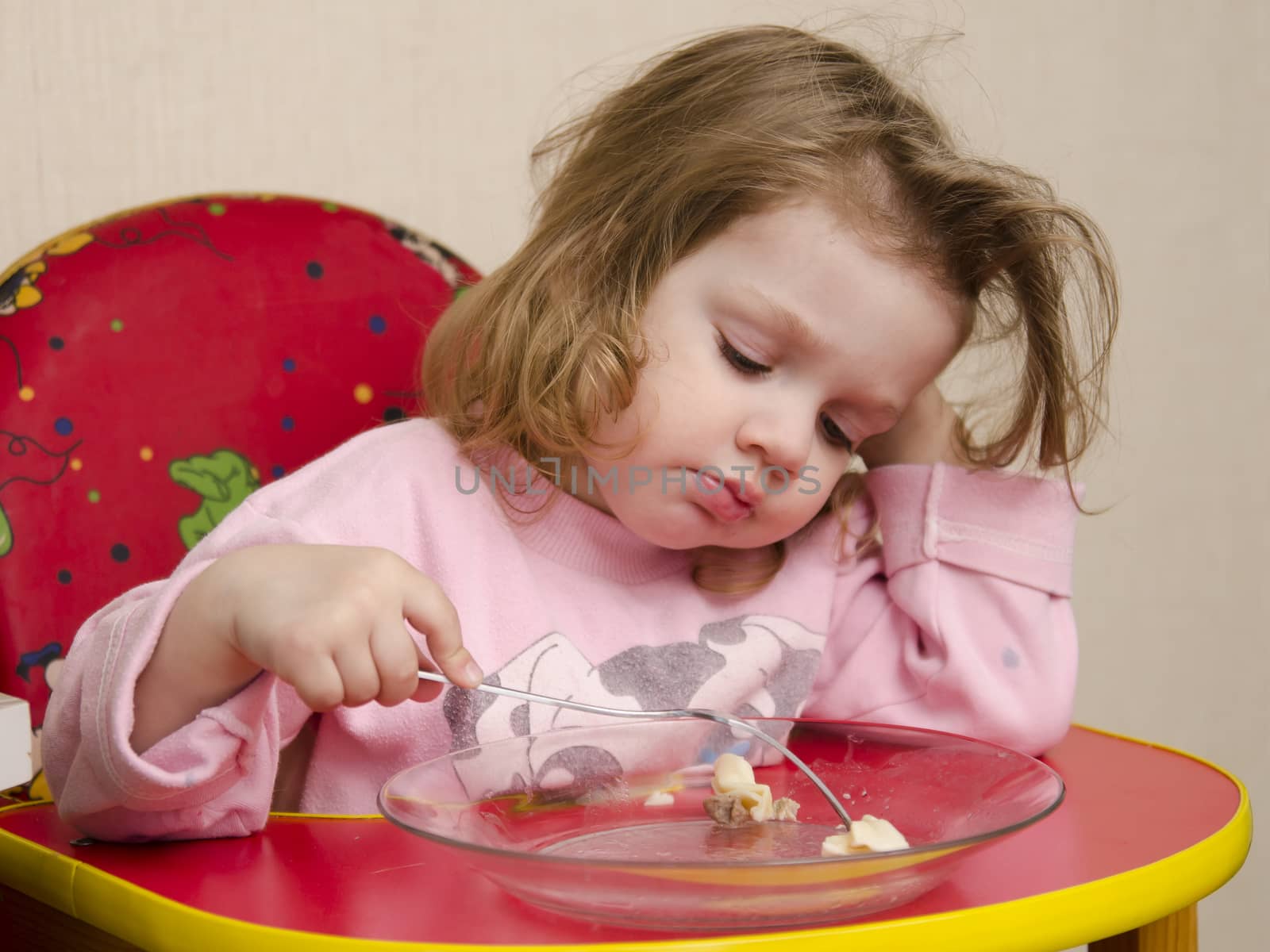 Two-year-old girl eats sitting at the table in the kitchen. Girl eats with a fork.
