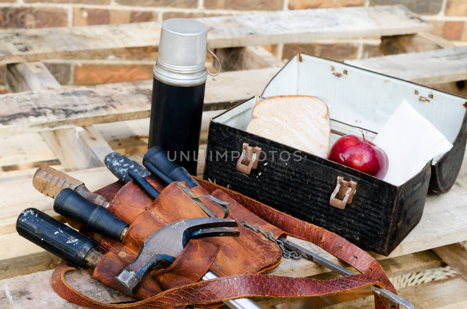 Lunch break at construction site with lunch pail, beverage container, apple, sandwich, napkin, tool belt, hammer, wrenches, screwdrivers on pallet with brick wall in background.