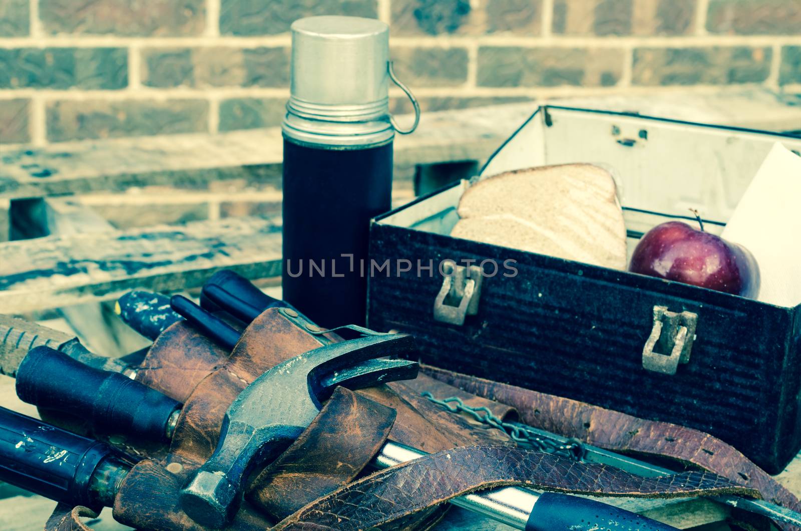 Worker's lunch break with lunch pail, beverage container, tool belt, hammer, screwdrivers,wrenches, apple, sandwich, and napkin on wooden pallet with brick wall in background. Image has been cross processed.