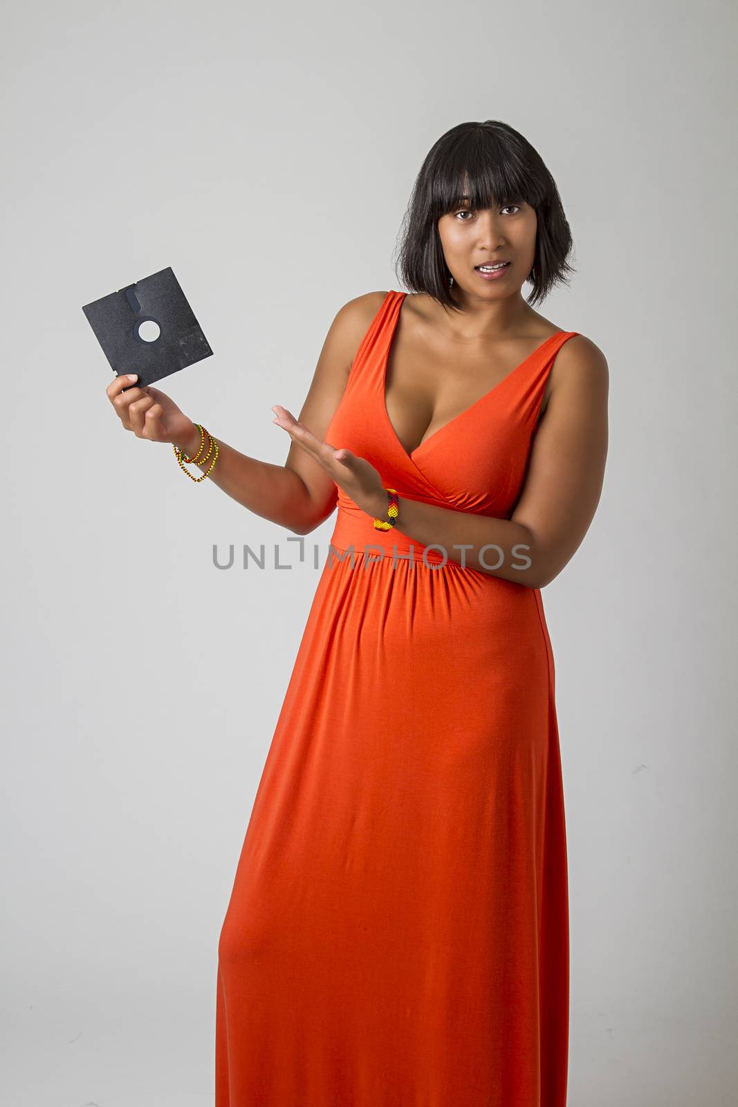 Sexy asian woman wearing a low cut orange dress, holding an old 5 in floppy disk, with questionning expression