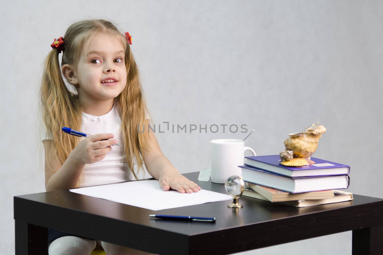 girl cheerfully and joyfully looks in the frame, sitting at the table. In one arm and the girls handle, which she is going to write on a sheet of paper. There are tombs of books, sheets of paper, a pen, glass globe, with a glass of tea, sea shell