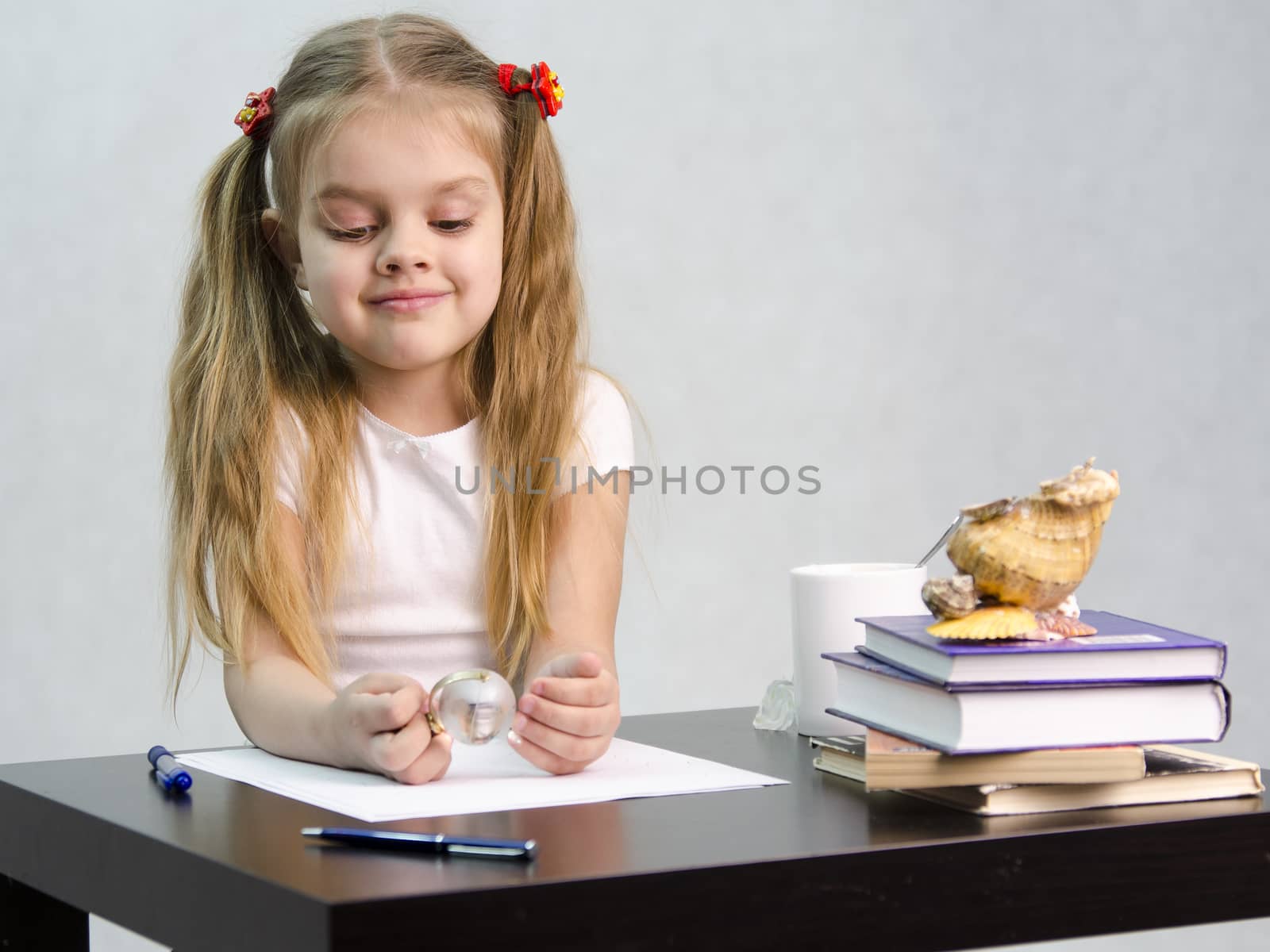 girl thoughtfully and effortlessly turns a small glass globe, sitting at the table. There are tombs of books, sheets of paper, a pen, glass globe, with a glass of tea, sea shell. The image of the writer.