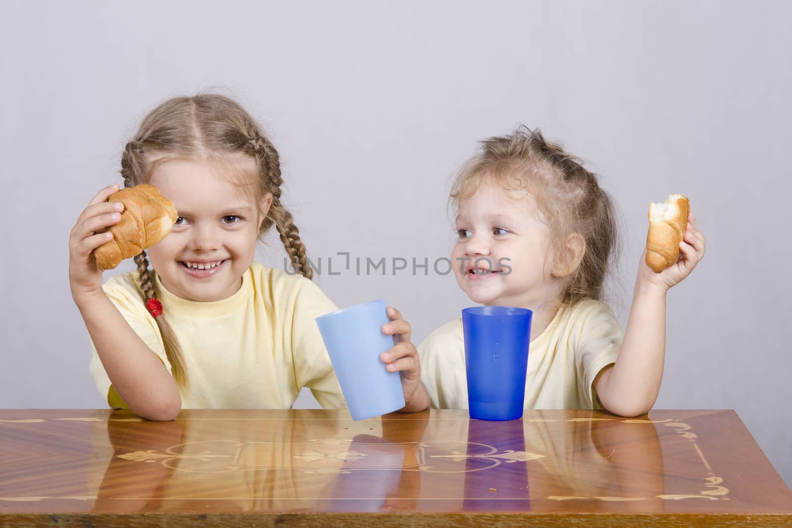 Two children sitting at the table, eating a muffin and drink of colored plastic cups