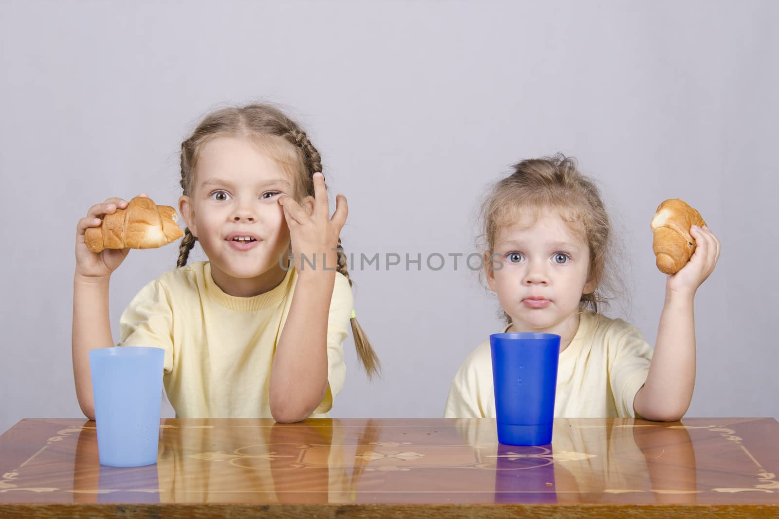 Two children sitting at the table, eating a muffin and drink of colored plastic cups