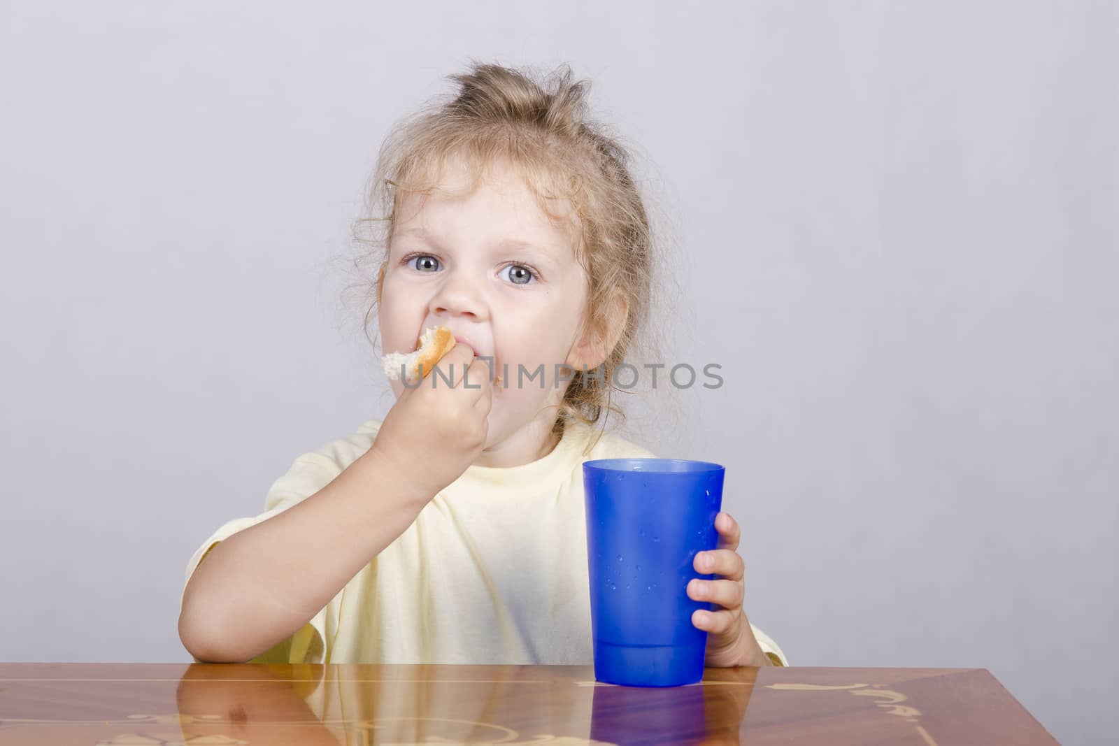 Two-year-old girl sitting at the table, eating a muffin and drinking from a plastic Cup