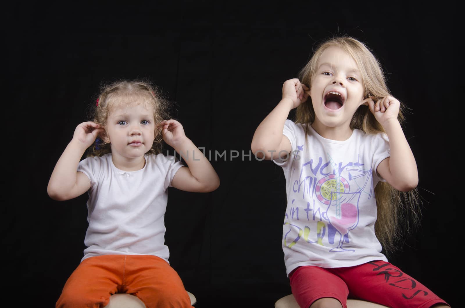 Portrait of the sisters - two little girls on the chairs. Girls fun smiling and looking in the frame. Girls hold hands behind your ears