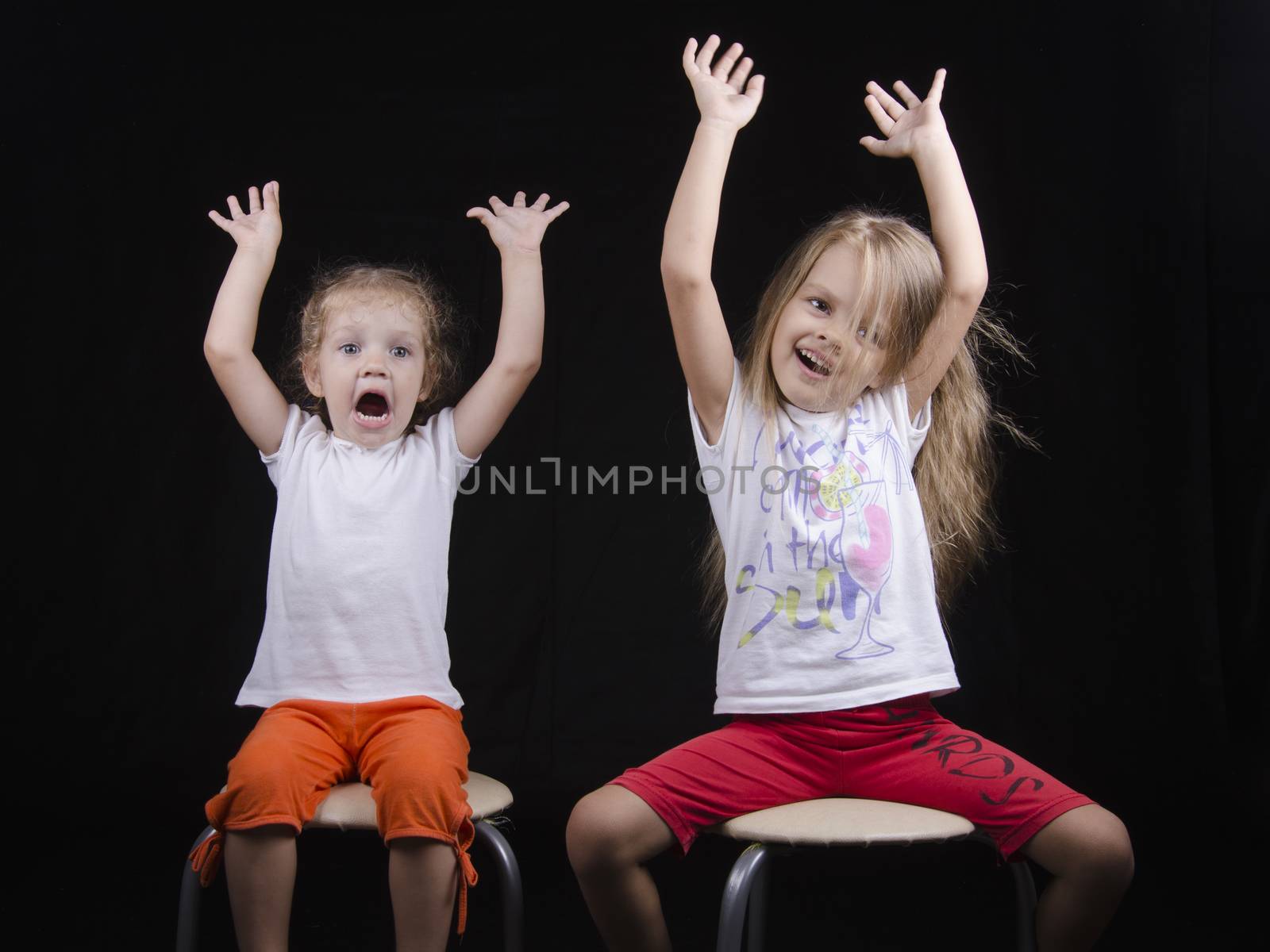 Portrait of the sisters - two little girls on the chairs. Girls fun smiling and looking in the frame. The girls raised their hands up.