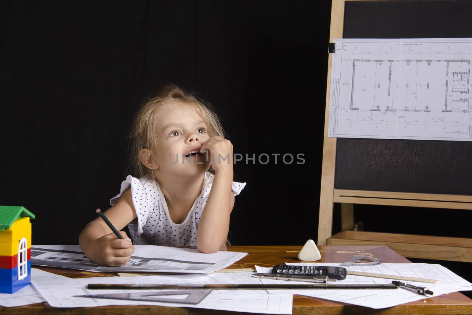 Four-year-old girl in the image of the architect. The girl sitting at the table with the drawings. In the background stands the Board with the drawing.