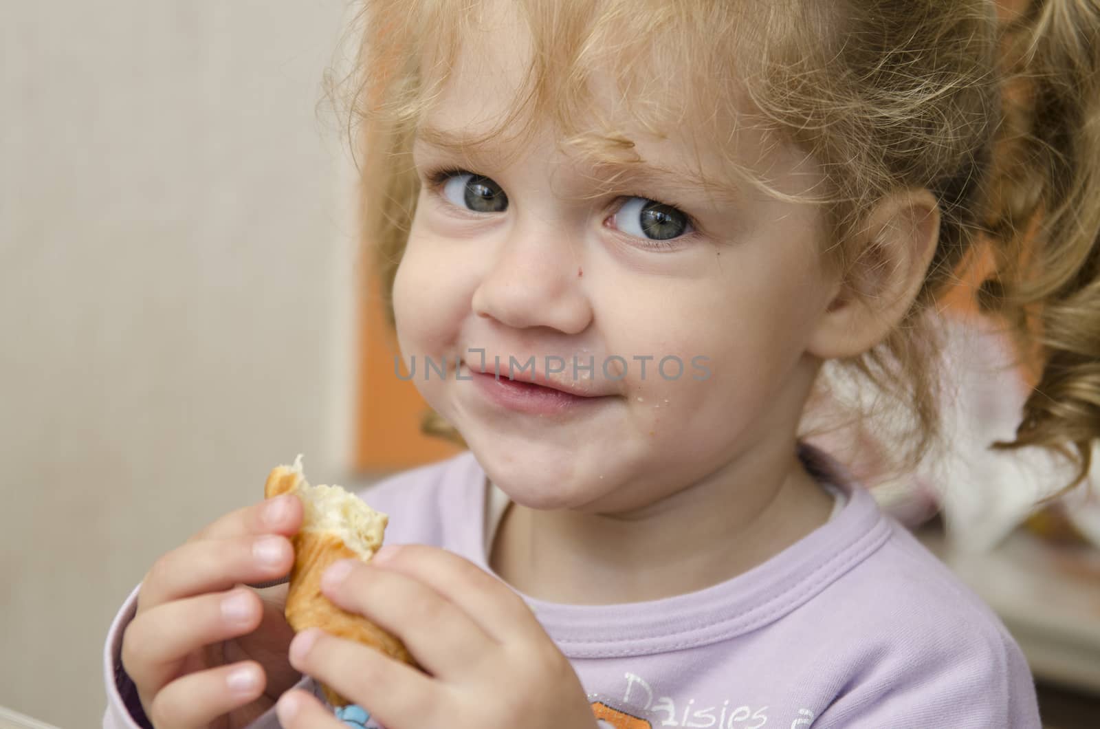 The little girl sits at a table and with enthusiasm eats a roll. Photo close up.