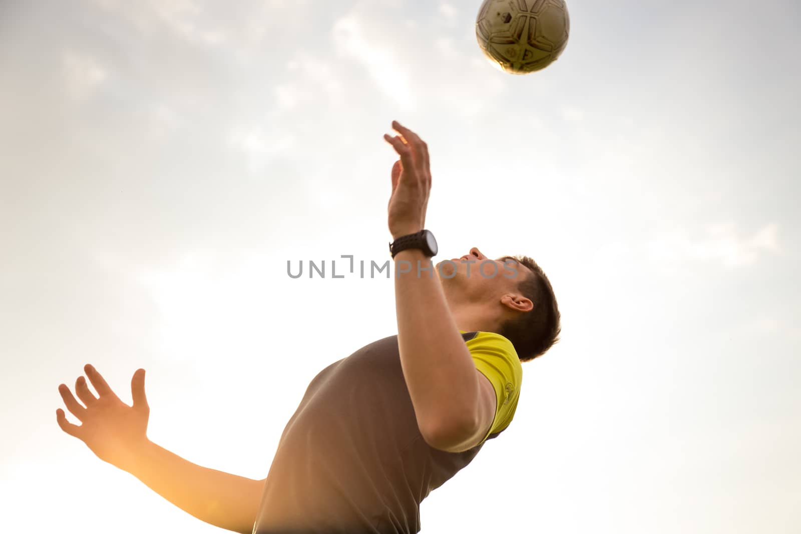 Young, male man playing soccer (lit by warm evening sunlight) heading the ball in the air