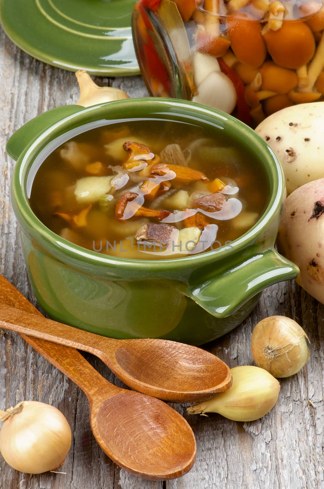 Delicious Vegetarian Soup with Chanterelle Mushrooms  in Green Pot with Raw Potato, Onion, Marinated Mushrooms and Wooden Spoons on closeup Rustic Wooden background