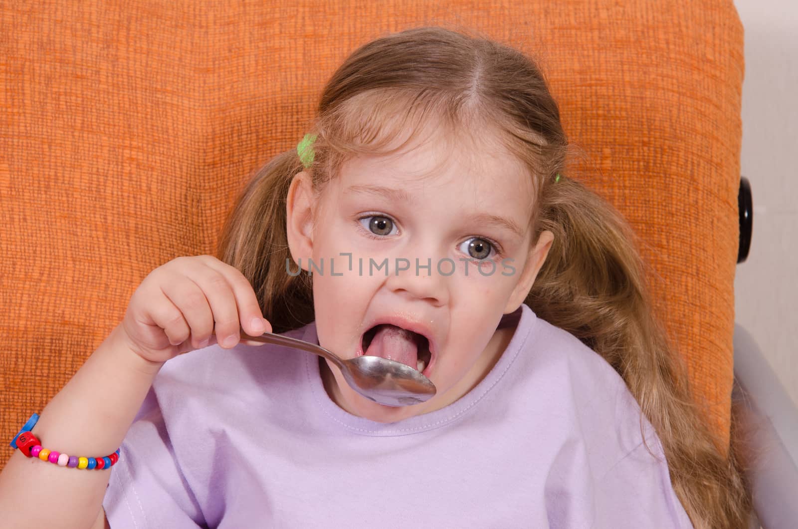 Three year old girl with pleasure and humor eats honey spoon sitting on the couch