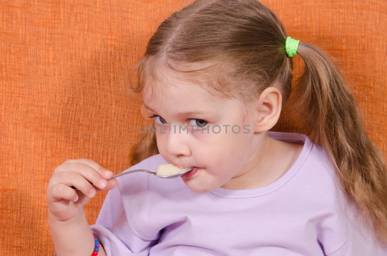 Three year old girl with pleasure and humor eats honey spoon sitting on the couch