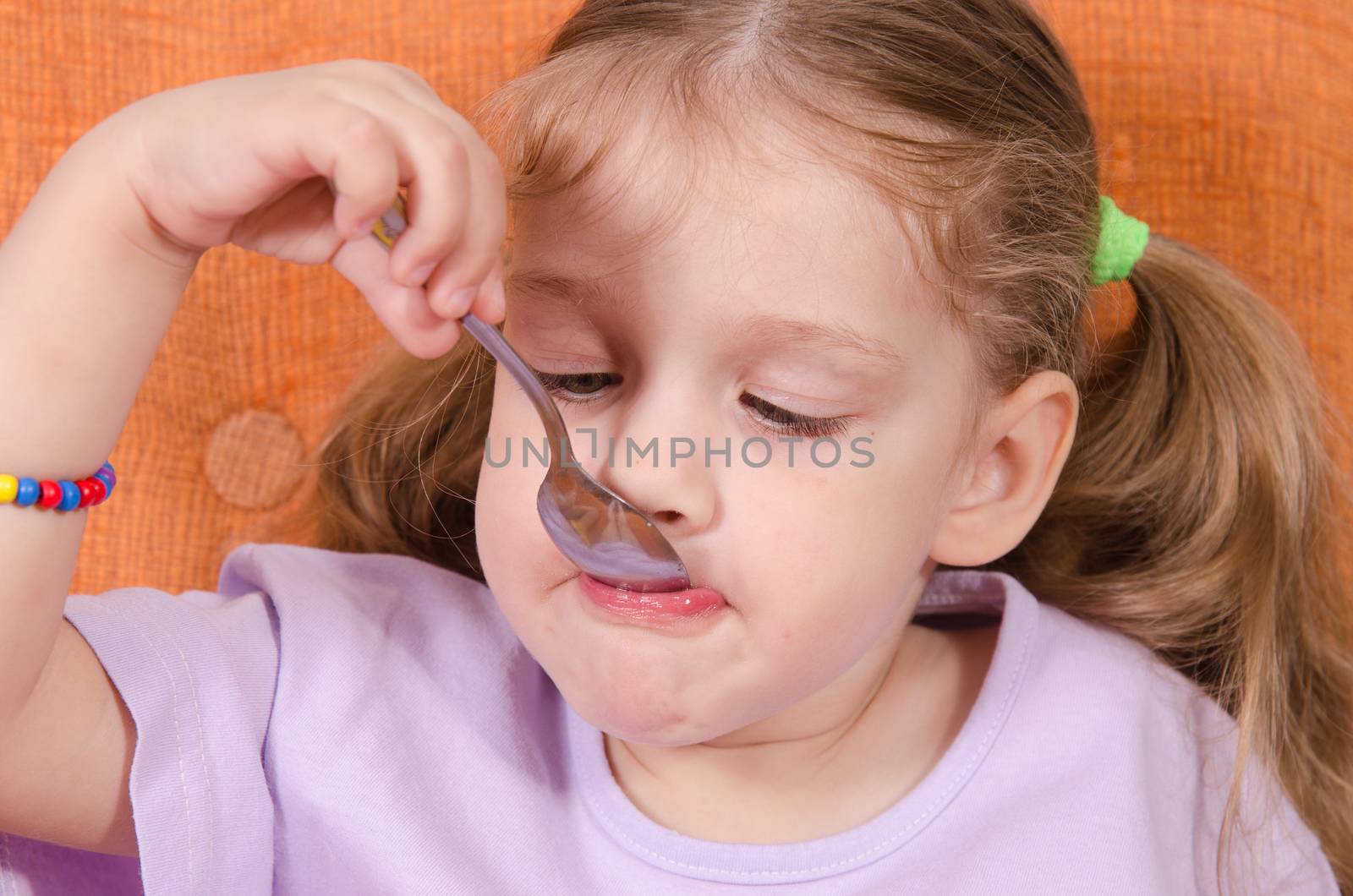 Three year old girl with pleasure and humor eats honey spoon sitting on the couch