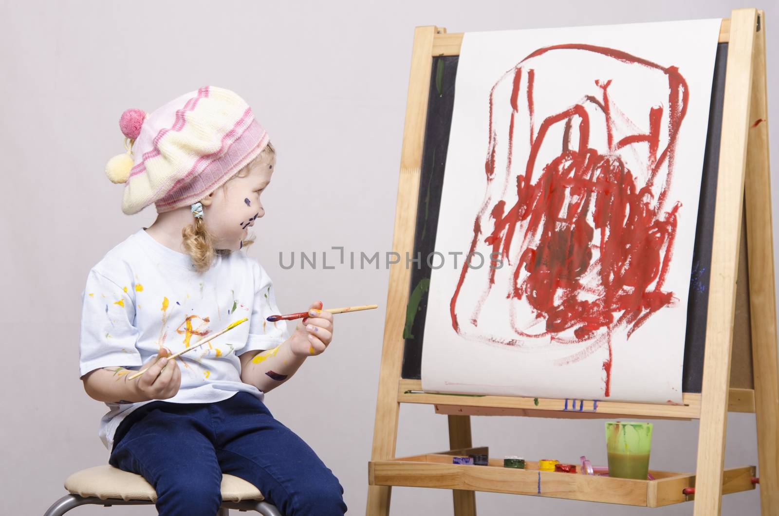 Portrait of a three-year old girl playing in the artist. Girl mud paints. Standing behind you easel