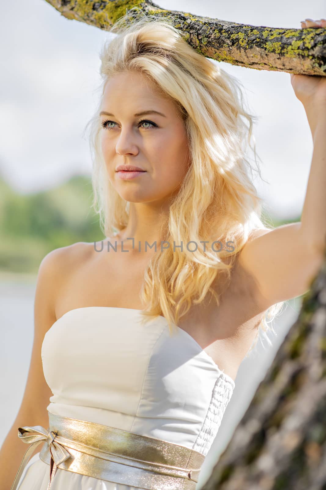Blond young woman standing under a tree at the edge of a lake