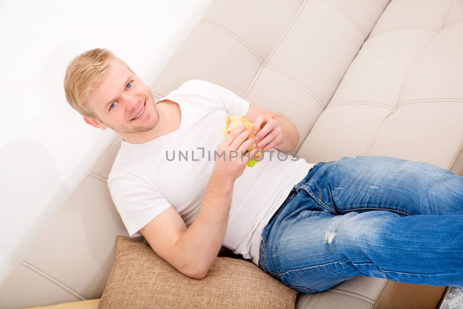 A young adult man eating a sandwich at home on the couch.
