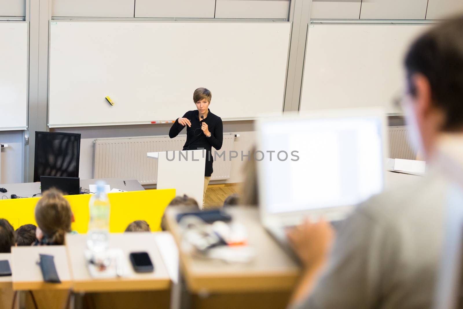 Speaker giving presentation in lecture hall at university. Participants listening to lecture and making notes.