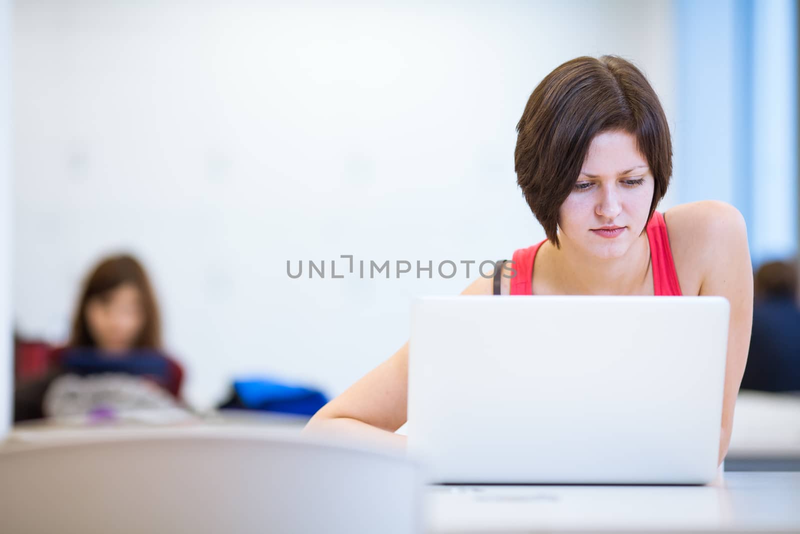 Pretty, young college student studying in the library/a study room at campus , using her modern laptop computer, writing notes, preparing for exams (shallow DOF)