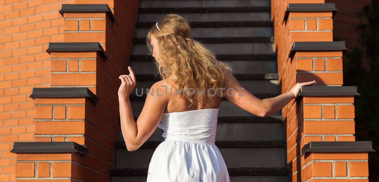 Coquettish young beautiful woman posing near the stairs at sunset. View from back